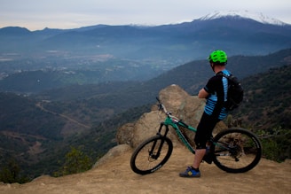 A cyclist wearing a green helmet and blue-striped jersey stands on a rocky ledge, looking out over a vast landscape. The view includes rolling hills and a distant snow-capped mountain under a cloudy sky. The mountain bike is positioned near the edge, suggesting a pause during a trail ride.