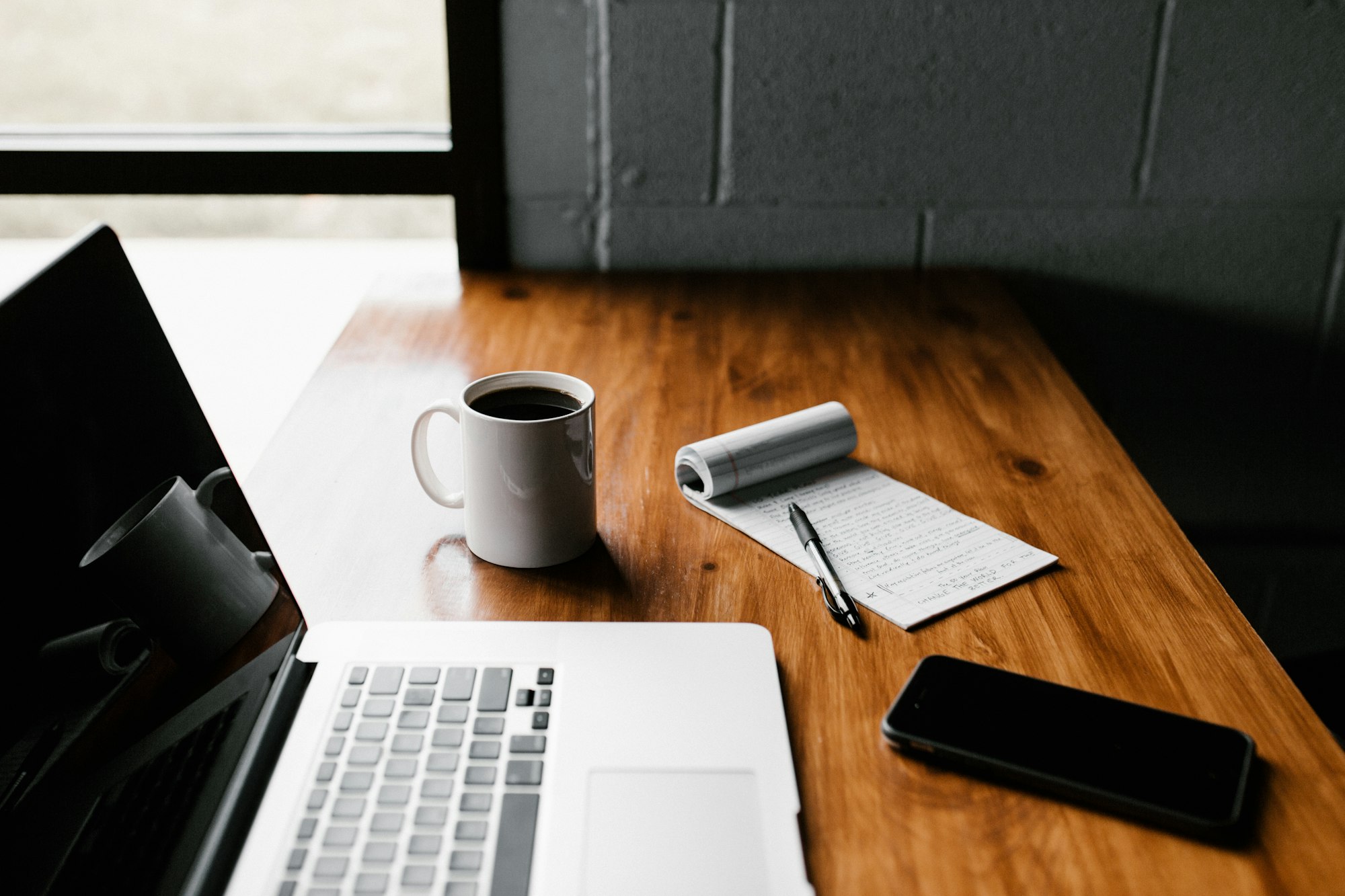 A laptop, iPhone, coffee, and notepad on a table.