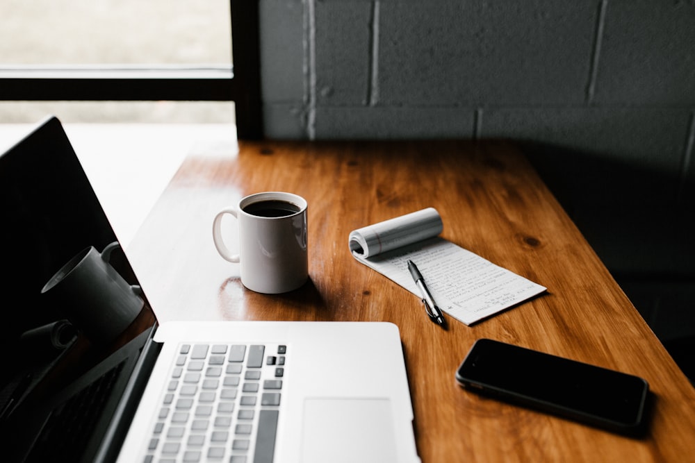 MacBook Pro, caneca de cerâmica branca e smartphone preto na mesa