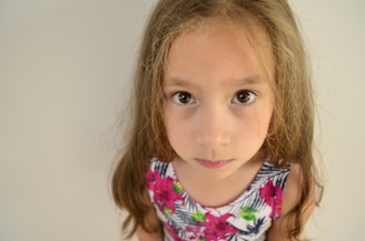 A young girl with long brown hair wearing a dress with a floral pattern. She is looking directly at the camera with a serious expression. The background is a plain light-colored wall.