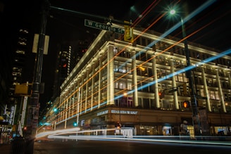 A large, illuminated building at night with streaks of light from passing vehicles in the foreground. The building, which appears to be a department store, has decorative lighting highlighting its architecture. There is a street sign indicating 'W Georgia St' and light trails creating a dynamic effect.