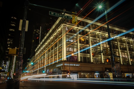 A large, illuminated building at night with streaks of light from passing vehicles in the foreground. The building, which appears to be a department store, has decorative lighting highlighting its architecture. There is a street sign indicating 'W Georgia St' and light trails creating a dynamic effect.