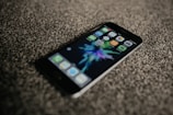A smartphone is lying on a textured carpeted surface. The screen displays colorful app icons and a vibrant, abstract background. The phone is oriented at a slight angle, with its top left corner closest to the viewer.