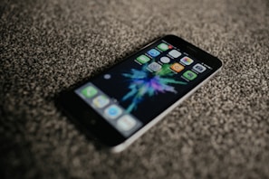 A smartphone is lying on a textured carpeted surface. The screen displays colorful app icons and a vibrant, abstract background. The phone is oriented at a slight angle, with its top left corner closest to the viewer.