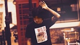 A person with dark hair and a tattooed arm is wearing a black t-shirt that has a graphic and the words 'WILD YOUTH' on it. They are positioned in front of a red telephone booth on a street. The background features a partially visible building and some street furniture.