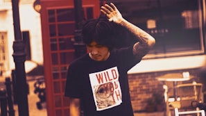 A person with dark hair and a tattooed arm is wearing a black t-shirt that has a graphic and the words 'WILD YOUTH' on it. They are positioned in front of a red telephone booth on a street. The background features a partially visible building and some street furniture.