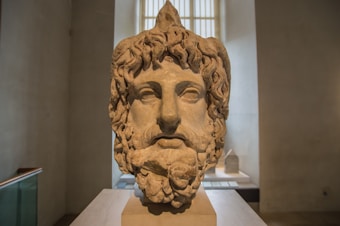 A large stone sculpture of a bearded man with curly hair, positioned in a museum setting. The sculpture exhibits intricate detailing in the hair and facial features, conveying a sense of antiquity. It is placed on a pedestal with soft, ambient lighting highlighting its textures against a neutral background.