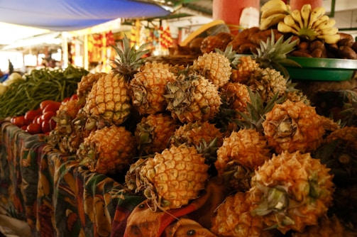 A vibrant market stall piled high with fresh pineapples, surrounded by other fruits and vegetables like bananas, tomatoes, and green beans. The tropical produce is displayed on a colorful patterned cloth, with a background that suggests a bustling, busy marketplace atmosphere.