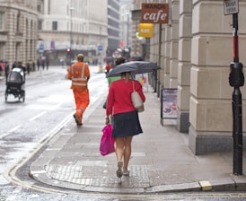 A rainy urban street scene features a person wearing a red jacket and carrying a pink shopping bag while holding an umbrella. The street is wet, and a worker in an orange uniform walks ahead. The scene includes outdoor signage for a cafe and cycling is restricted as indicated by a road sign.