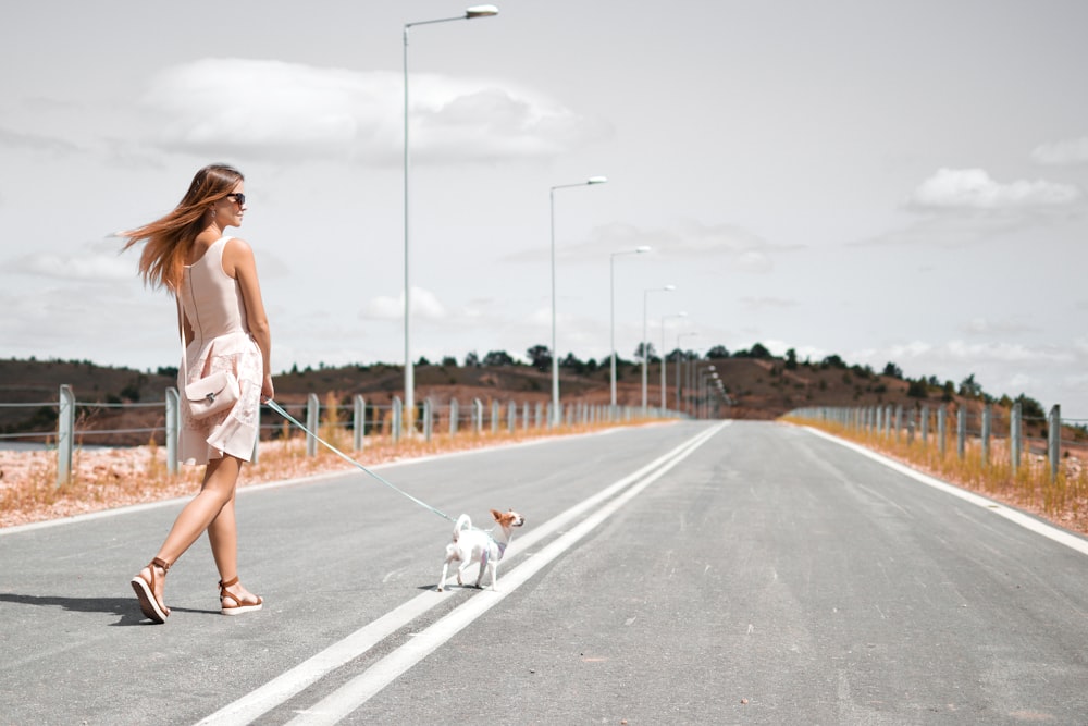 흰색 민소매 드레스를 입은 여자가 낮에 아스팔트 도로를 걷는 강아지의 하네스를 들고 있습니다.
