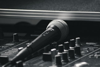 A black and silver microphone resting on a sound mixing console. The image emphasizes the equipment's details, with various knobs and sliders visible in the foreground. The scene has a professional studio vibe, suggesting a focus on audio production.