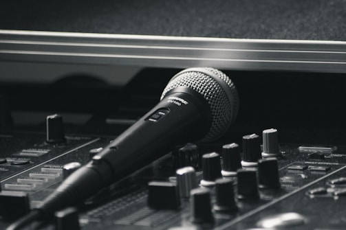 A black and silver microphone resting on a sound mixing console. The image emphasizes the equipment's details, with various knobs and sliders visible in the foreground. The scene has a professional studio vibe, suggesting a focus on audio production.