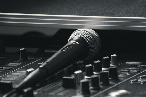A black and silver microphone resting on a sound mixing console. The image emphasizes the equipment's details, with various knobs and sliders visible in the foreground. The scene has a professional studio vibe, suggesting a focus on audio production.
