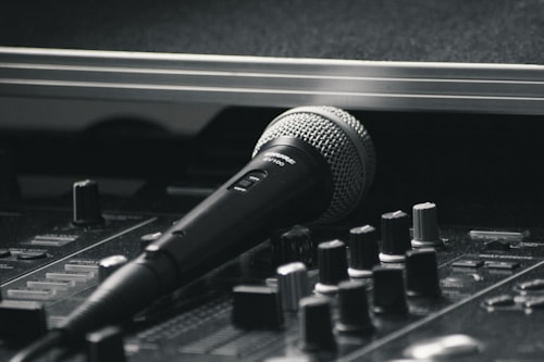 A black and silver microphone resting on a sound mixing console. The image emphasizes the equipment's details, with various knobs and sliders visible in the foreground. The scene has a professional studio vibe, suggesting a focus on audio production.
