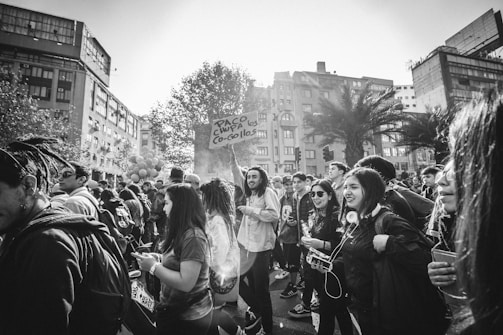 A group of people gather in an urban area, with some holding signs and others engaging with their phones or each other. The scene appears lively, with a variety of individuals, including those wearing backpacks and sunglasses. Overhead, trees and tall buildings frame the location, suggesting a busy city environment.
