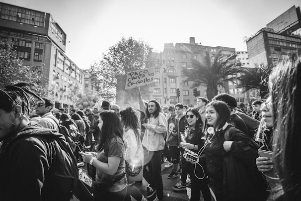 A group of people gather in an urban area, with some holding signs and others engaging with their phones or each other. The scene appears lively, with a variety of individuals, including those wearing backpacks and sunglasses. Overhead, trees and tall buildings frame the location, suggesting a busy city environment.