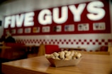 A tray of peanuts sits on a wooden table within a fast-food restaurant setting. The background features a large red and white sign with illuminated letters, and the interior includes red accents and wooden furniture.