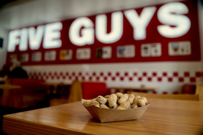 A tray of peanuts sits on a wooden table within a fast-food restaurant setting. The background features a large red and white sign with illuminated letters, and the interior includes red accents and wooden furniture.