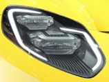 A close-up of a car headlight featuring a sleek and modern design with LED light strips and a glossy surface. The headlight is set against a bright yellow car body with reflective highlights.