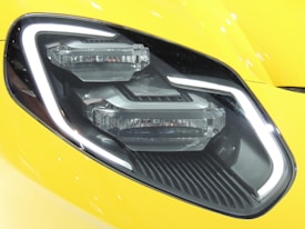 A close-up of a car headlight featuring a sleek and modern design with LED light strips and a glossy surface. The headlight is set against a bright yellow car body with reflective highlights.