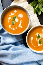 Two bowls of creamy orange soup garnished with a swirl of cream and fresh basil leaves are presented. The background includes a rustic note and fresh basil leaves on the side. A light blue cloth and dark tabletop frame the bowls.