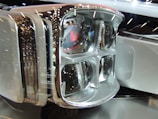 Close-up of a modern, sleek headlight assembly with multiple bulbs and a chrome finish. The reflective surface and intricate detailing give it a futuristic appearance, highlighted by surrounding ambient light reflections.
