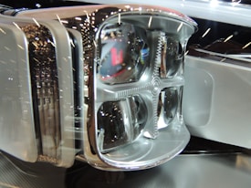 Close-up of a modern, sleek headlight assembly with multiple bulbs and a chrome finish. The reflective surface and intricate detailing give it a futuristic appearance, highlighted by surrounding ambient light reflections.