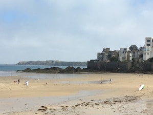 A sandy beach with scattered people enjoying leisure activities. A row of houses lines the shore, with rocky outcrops and an island visible in the distance under a partly cloudy sky.