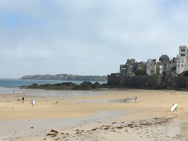 A sandy beach with scattered people enjoying leisure activities. A row of houses lines the shore, with rocky outcrops and an island visible in the distance under a partly cloudy sky.