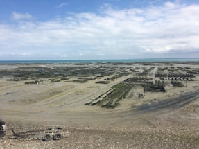 A coastal area with large, organized structures that appear to be aquaculture farms, possibly oyster or mussel beds. The scene is expansive, stretching towards the horizon, under a cloudy sky with patches of blue.