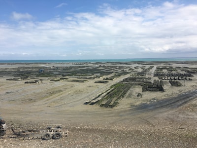 A coastal area with large, organized structures that appear to be aquaculture farms, possibly oyster or mussel beds. The scene is expansive, stretching towards the horizon, under a cloudy sky with patches of blue.
