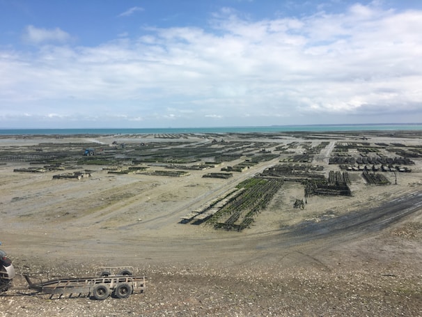 A coastal area with large, organized structures that appear to be aquaculture farms, possibly oyster or mussel beds. The scene is expansive, stretching towards the horizon, under a cloudy sky with patches of blue.