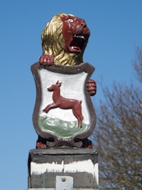 A detailed lion statue design features a fierce, open-mouthed lion with a red face and yellow mane, grasping a shield. The shield depicts a red deer leaping on a green and white background.