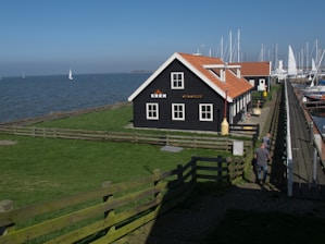 A black wooden building with a red-tiled roof is situated near a body of water with sailboats in the distance. The building has 'KNRM' written on it. A grassy area with a wooden fence surrounds the building. The scene includes a clear blue sky, and a few people walking along a path adjacent to a marina filled with more sailboats.