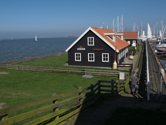 A black wooden building with a red-tiled roof is situated near a body of water with sailboats in the distance. The building has 'KNRM' written on it. A grassy area with a wooden fence surrounds the building. The scene includes a clear blue sky, and a few people walking along a path adjacent to a marina filled with more sailboats.