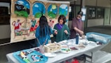 Three people are engaged in an art project, working on a mural with a colorful scene depicting figures dancing and landscapes. Several art supplies, including paint tubes, brushes, and plates with mixed colors, are spread across a table. The mural features vibrant blues, greens, and earthy tones.