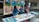 Three people are engaged in an art project, working on a mural with a colorful scene depicting figures dancing and landscapes. Several art supplies, including paint tubes, brushes, and plates with mixed colors, are spread across a table. The mural features vibrant blues, greens, and earthy tones.