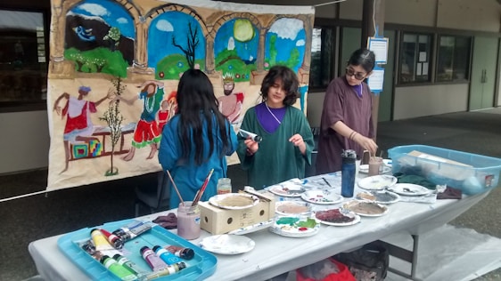 Three people are engaged in an art project, working on a mural with a colorful scene depicting figures dancing and landscapes. Several art supplies, including paint tubes, brushes, and plates with mixed colors, are spread across a table. The mural features vibrant blues, greens, and earthy tones.