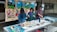 Three people are engaged in an art project, working on a mural with a colorful scene depicting figures dancing and landscapes. Several art supplies, including paint tubes, brushes, and plates with mixed colors, are spread across a table. The mural features vibrant blues, greens, and earthy tones.