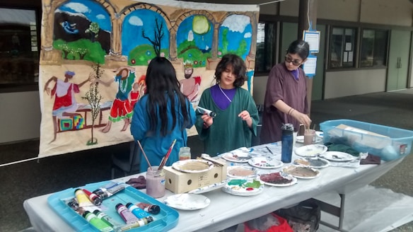 Three people are engaged in an art project, working on a mural with a colorful scene depicting figures dancing and landscapes. Several art supplies, including paint tubes, brushes, and plates with mixed colors, are spread across a table. The mural features vibrant blues, greens, and earthy tones.
