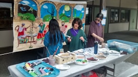 Three people are engaged in an art project, working on a mural with a colorful scene depicting figures dancing and landscapes. Several art supplies, including paint tubes, brushes, and plates with mixed colors, are spread across a table. The mural features vibrant blues, greens, and earthy tones.