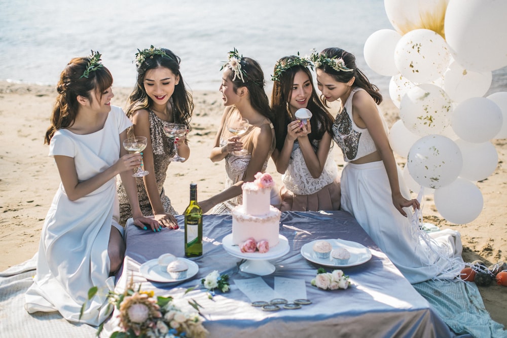 Um grupo de jovens mulheres asiáticas elegantemente vestidas ao lado de uma mesa com bolo, vinho e cupcakes