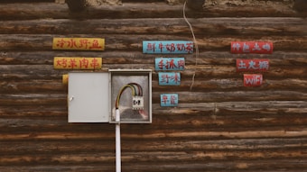 A rustic wooden wall features an open electrical panel with exposed wiring. Surrounding the panel are colorful signs with text in a language likely to be Chinese. The signs are mounted on the wall in a scattered arrangement, offering a contrast between modern technology and traditional aesthetics.