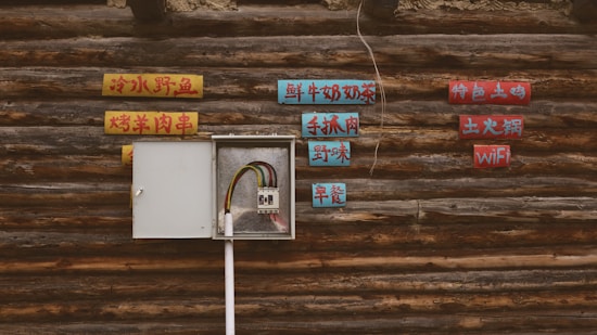 A rustic wooden wall features an open electrical panel with exposed wiring. Surrounding the panel are colorful signs with text in a language likely to be Chinese. The signs are mounted on the wall in a scattered arrangement, offering a contrast between modern technology and traditional aesthetics.