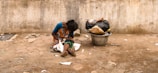 A person sits on the ground against a rough, weathered wall. The person is dressed in a colorful saree and appears to be looking down. Beside them, there is a large metal container filled with bags and trash. The ground is littered with scattered debris, and the overall scene suggests a setting of poverty and hardship.