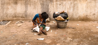 A person sits on the ground against a rough, weathered wall. The person is dressed in a colorful saree and appears to be looking down. Beside them, there is a large metal container filled with bags and trash. The ground is littered with scattered debris, and the overall scene suggests a setting of poverty and hardship.