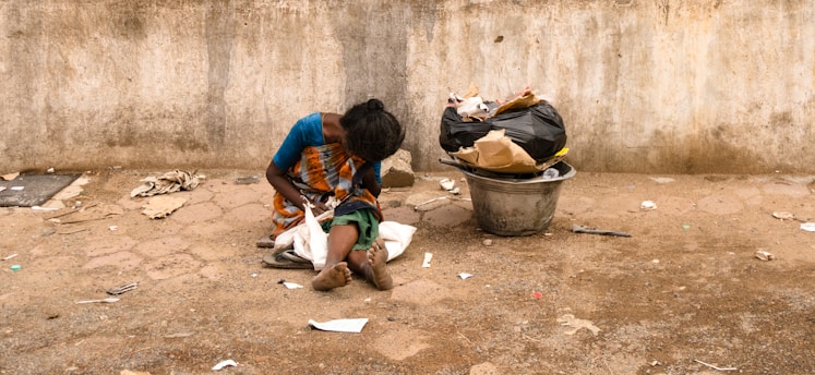 A person sits on the ground against a rough, weathered wall. The person is dressed in a colorful saree and appears to be looking down. Beside them, there is a large metal container filled with bags and trash. The ground is littered with scattered debris, and the overall scene suggests a setting of poverty and hardship.