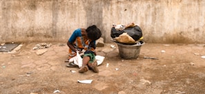 A person sits on the ground against a rough, weathered wall. The person is dressed in a colorful saree and appears to be looking down. Beside them, there is a large metal container filled with bags and trash. The ground is littered with scattered debris, and the overall scene suggests a setting of poverty and hardship.