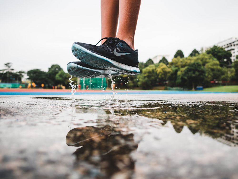 person wearing pair of gray-and-white Nike running shoes jumping on gray concrete floor with water during daytime