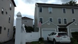A residential area with a three-story house featuring multiple windows and a garage, alongside a white fence. An SUV is parked in front of the garage, and an American flag is mounted near the entrance. The sky is overcast with clouds.